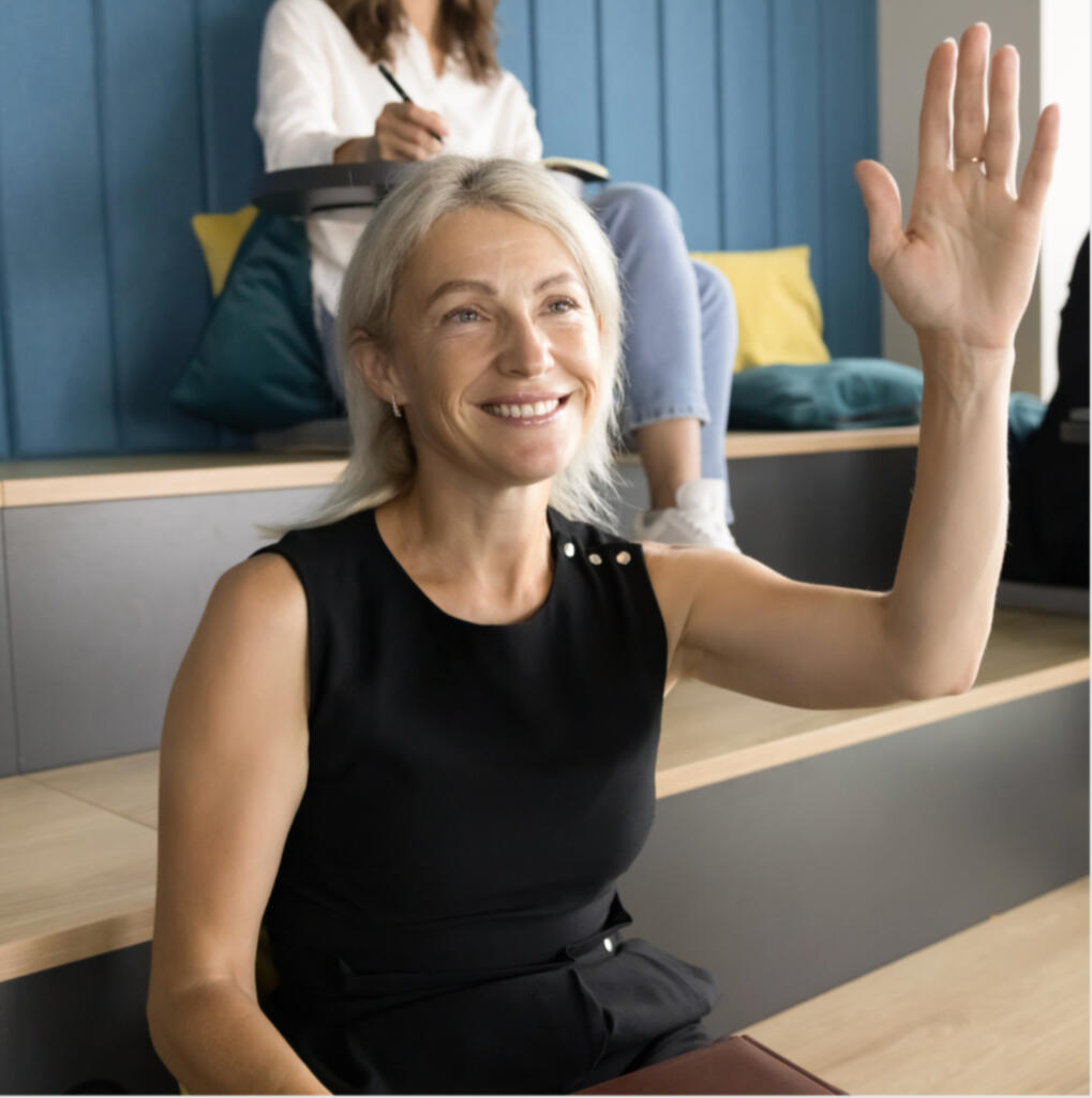 a confident smiling older woman with her hand raised seated in a black dress top